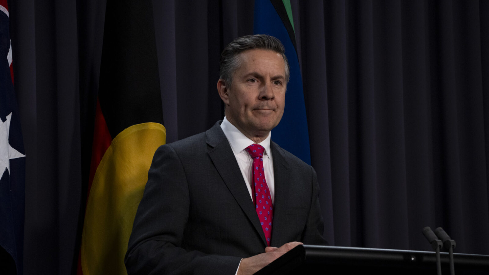 Mark Butler, a white man with a dark suit and red tie, stands speaking at a podium