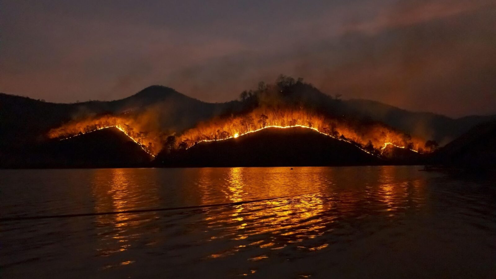 A distant image of a hill burning, with water in the foreground.