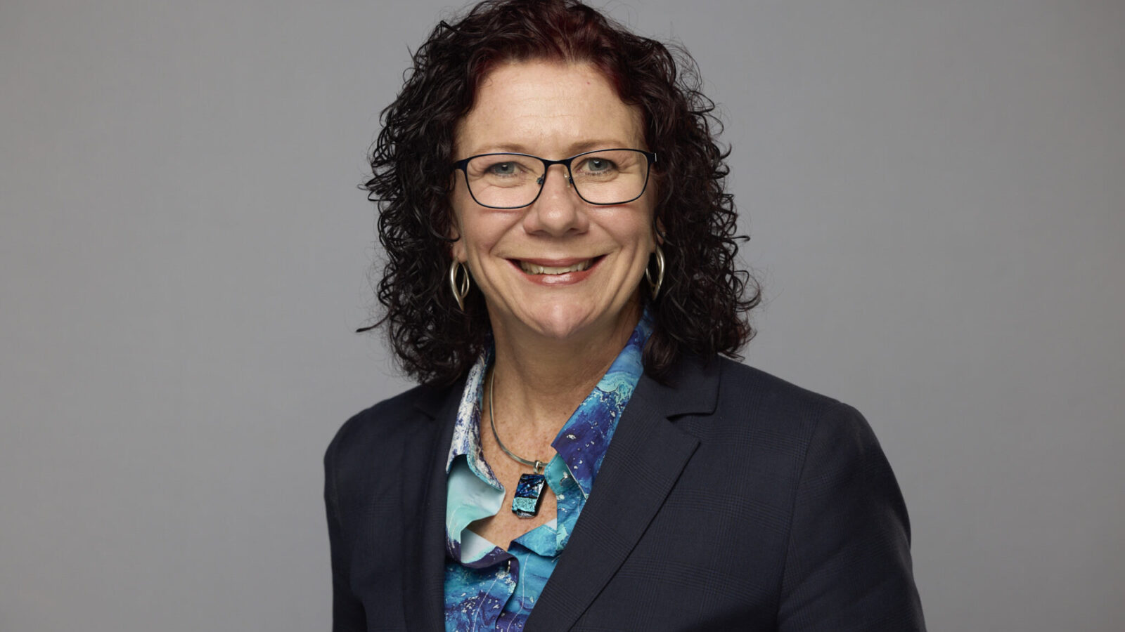 A photo of Jodie Griffiths-Cook, smiling with dark hair and thing dark glasses. She is wearing a blue top and dark jacket.