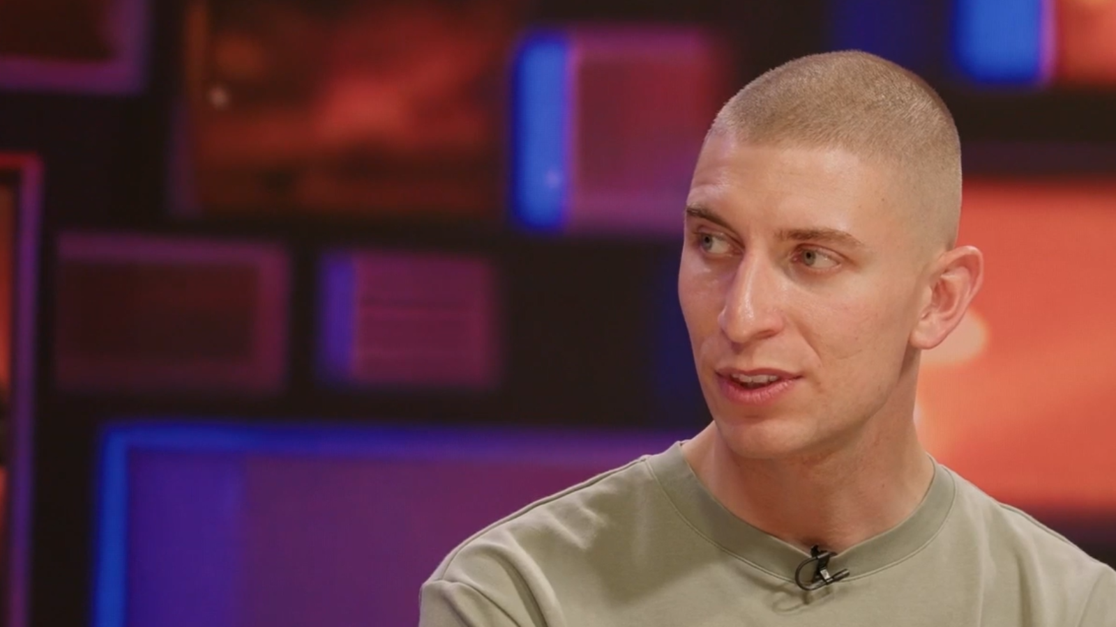 Jacob Darkin, sitting a the Reframed desk. They have a shaved head and are wearing a khaki tshirt.