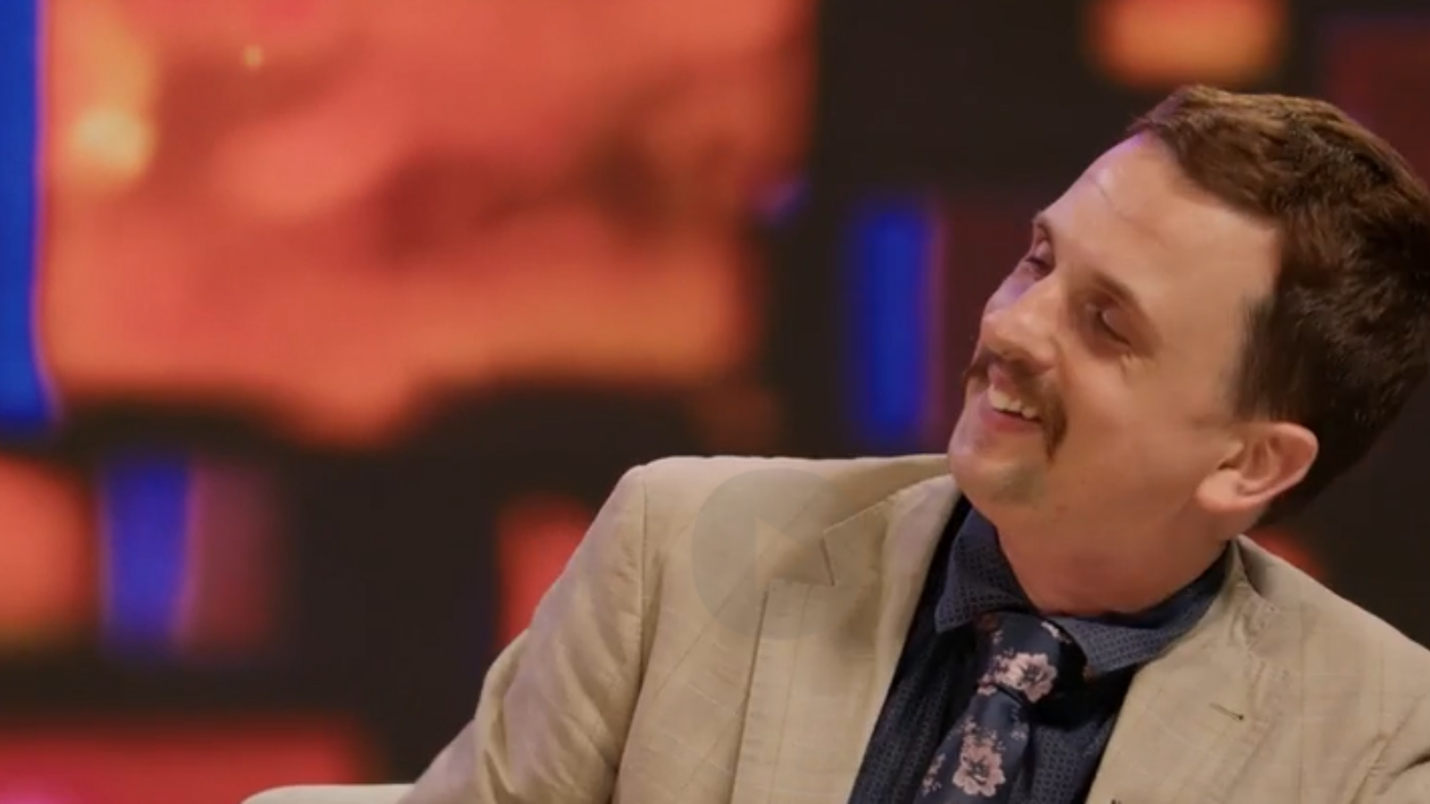 John is wearing a tan suit jacket with a dark shirt and tie, is leaning to his left and smiling