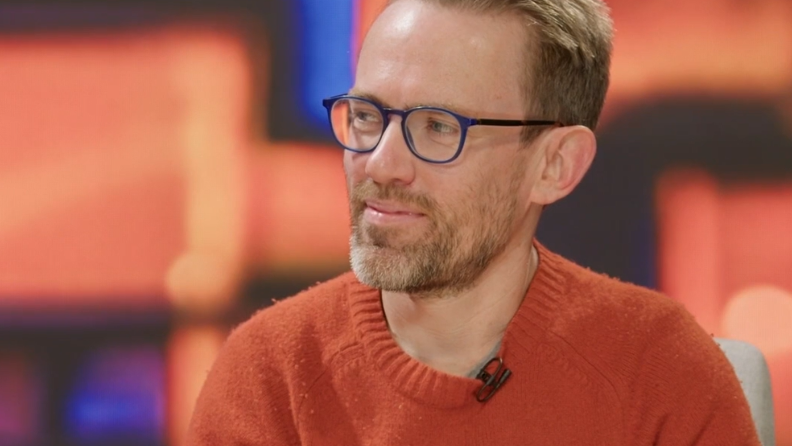 Sam looks to his right at the hosts, he is wearing an orange jumper with a microphone on it