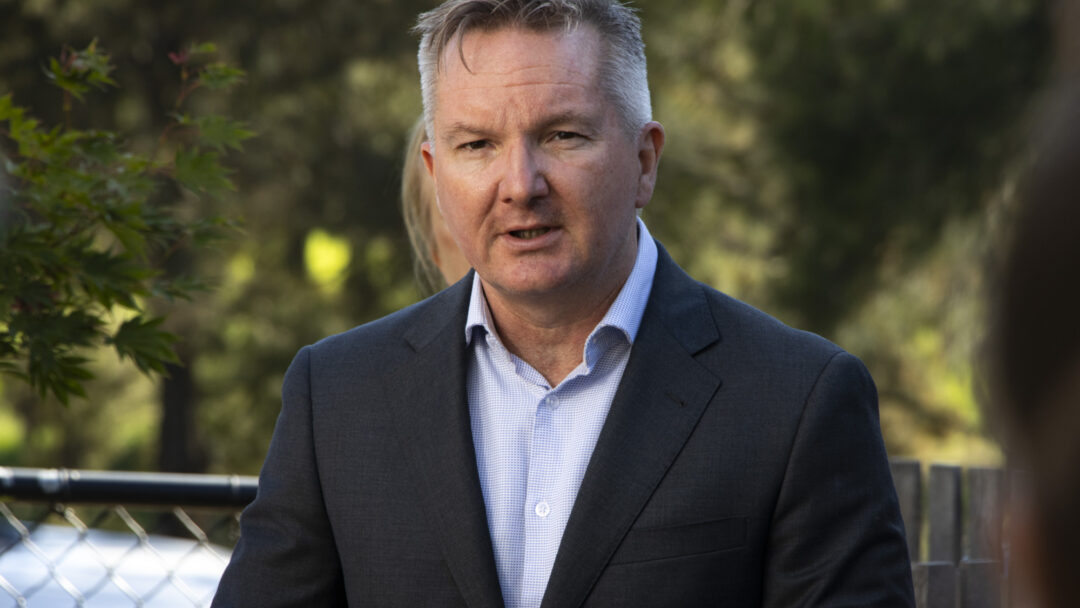Minister Chris Bowen speaking outdoors. He is wearing a light blue collared shirt and a dark jacket.