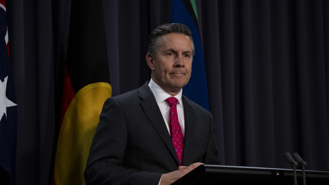 Mark Butler, a white man with a dark suit and red tie, stands speaking at a podium