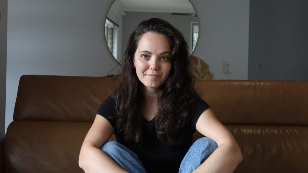 Dr Kristy Sommer, looking directly at the camera. They are wearing a black top and jeans and have long dark hair.