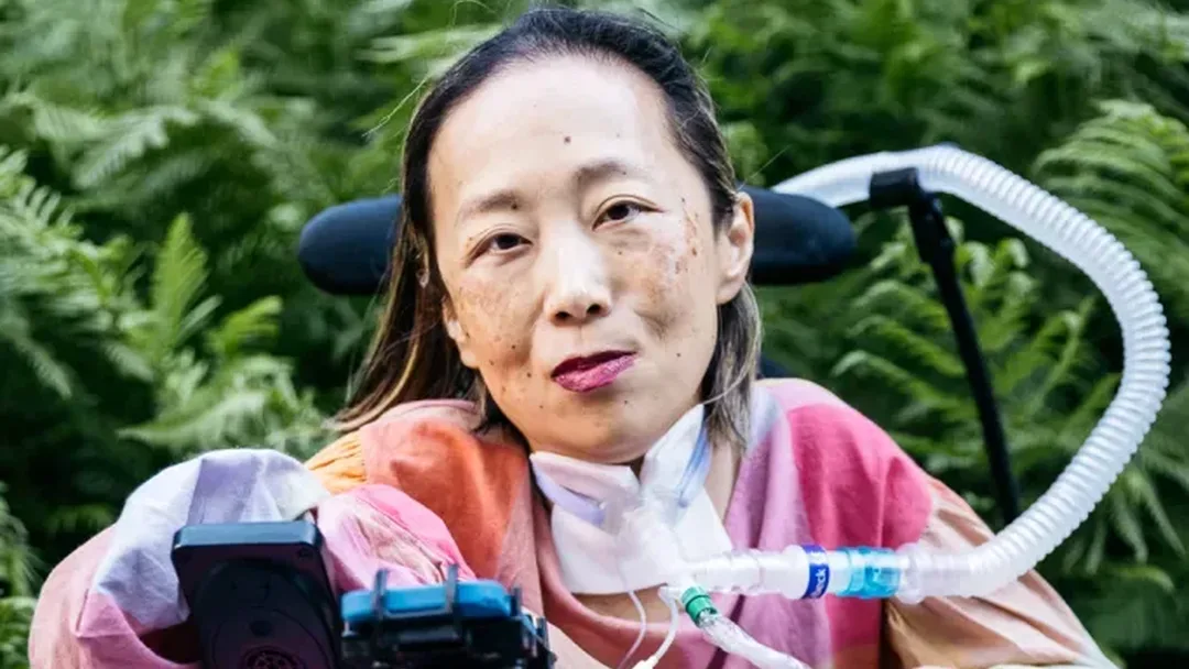 Alice Wong, Asian American woman in a wheelchair with a tracheostomy at her neck connected to a ventilator. She’s wearing a pink plaid shirt, pink pants, and a magenta lip color. She is smiling and behind her are a bunch of large fern-like plants. Photo credit: Allison Busch Photography.