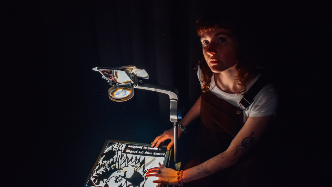 Bria McCarthy sits in a dark room beside an overhead projector. Her head is tilted upward, as if looking toward the projection. On the projector’s glass surface, Bria’s hands hold down a hand-drawn slide. The bright light from the projector partially illuminates her face and arms, while the rest of the scene remains in shadow