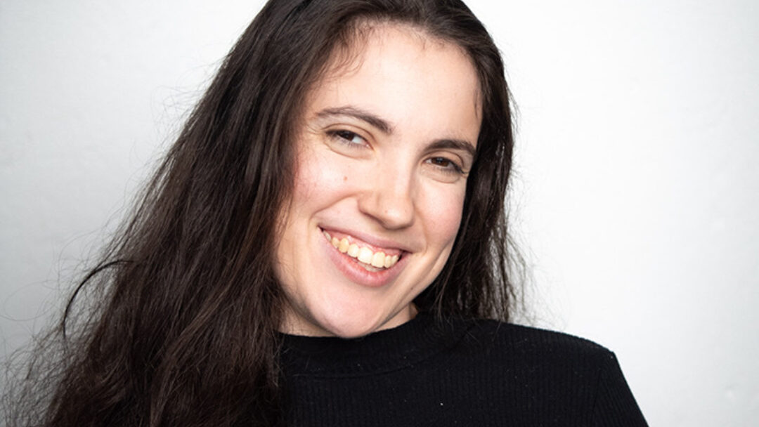 Hannah Diviney smiles at the camera. She has long dark hair and is wearing a black long sleeved shirt.
