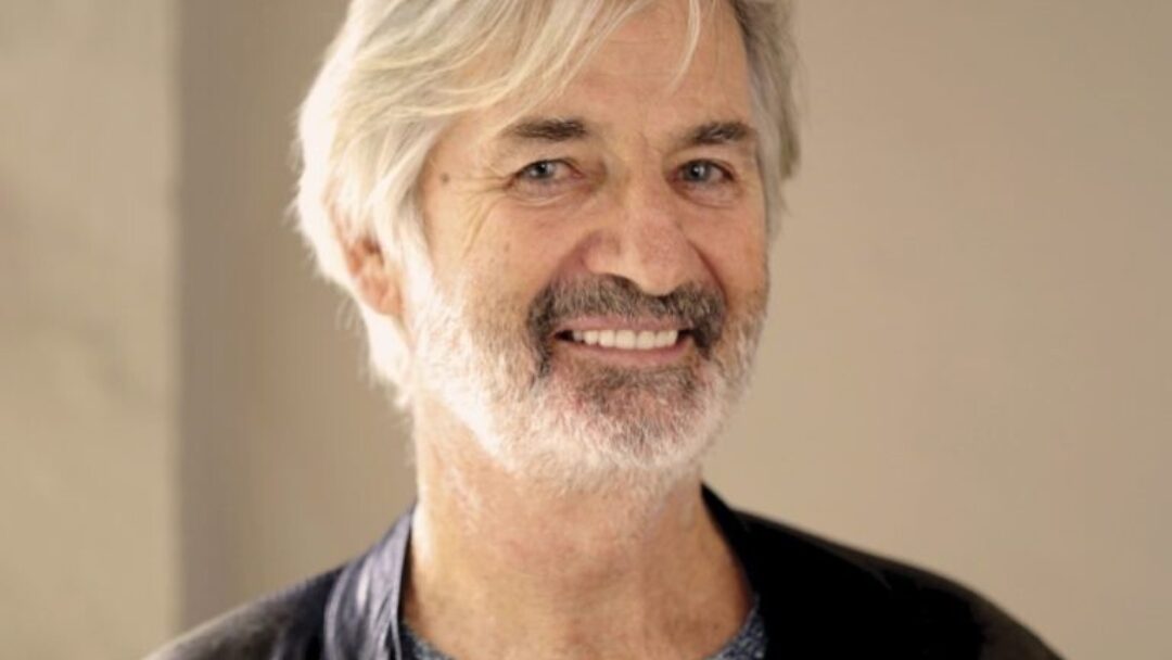 A smiling headshot of actor John Jarrett, with a light beard and grey hair