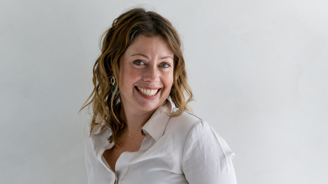 Michelle Olaithe, a smiling white woman who is wearing a white shirt and grey trousers