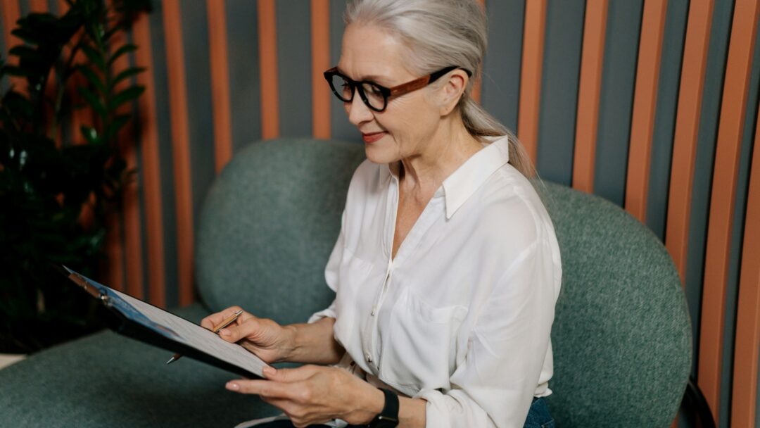 A person in a white shirt with grey hair and black glasses looks at documents they are holding