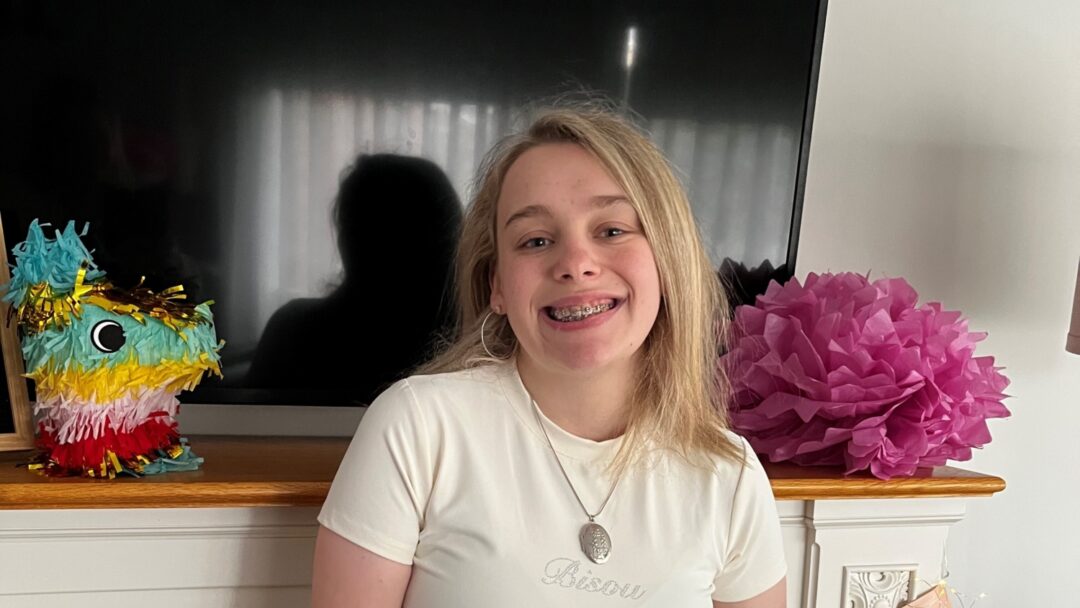 Realle Northe standing by a tv screen. She is wearing a white shirt and blue jeans and smiling at the camera.