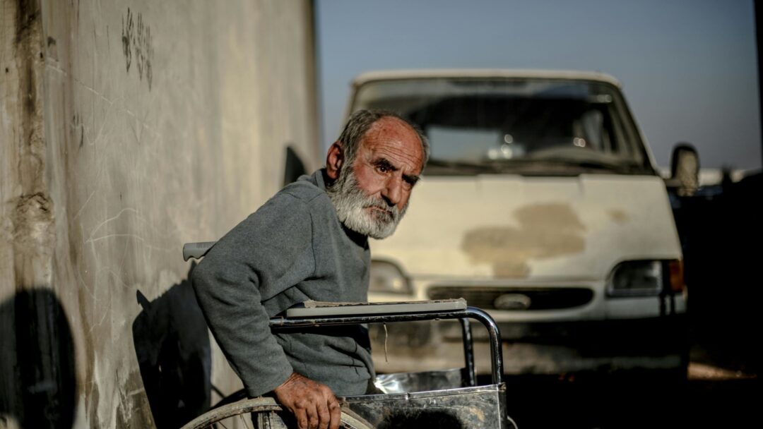 An older bearded man sits in an old manual wheelchair.