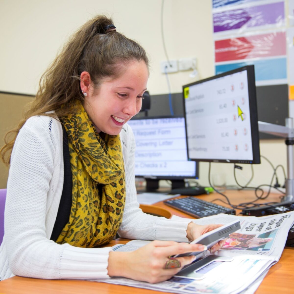 A woman sitting at an office desk is using a magnifying device to read a broadsheet newspaper.