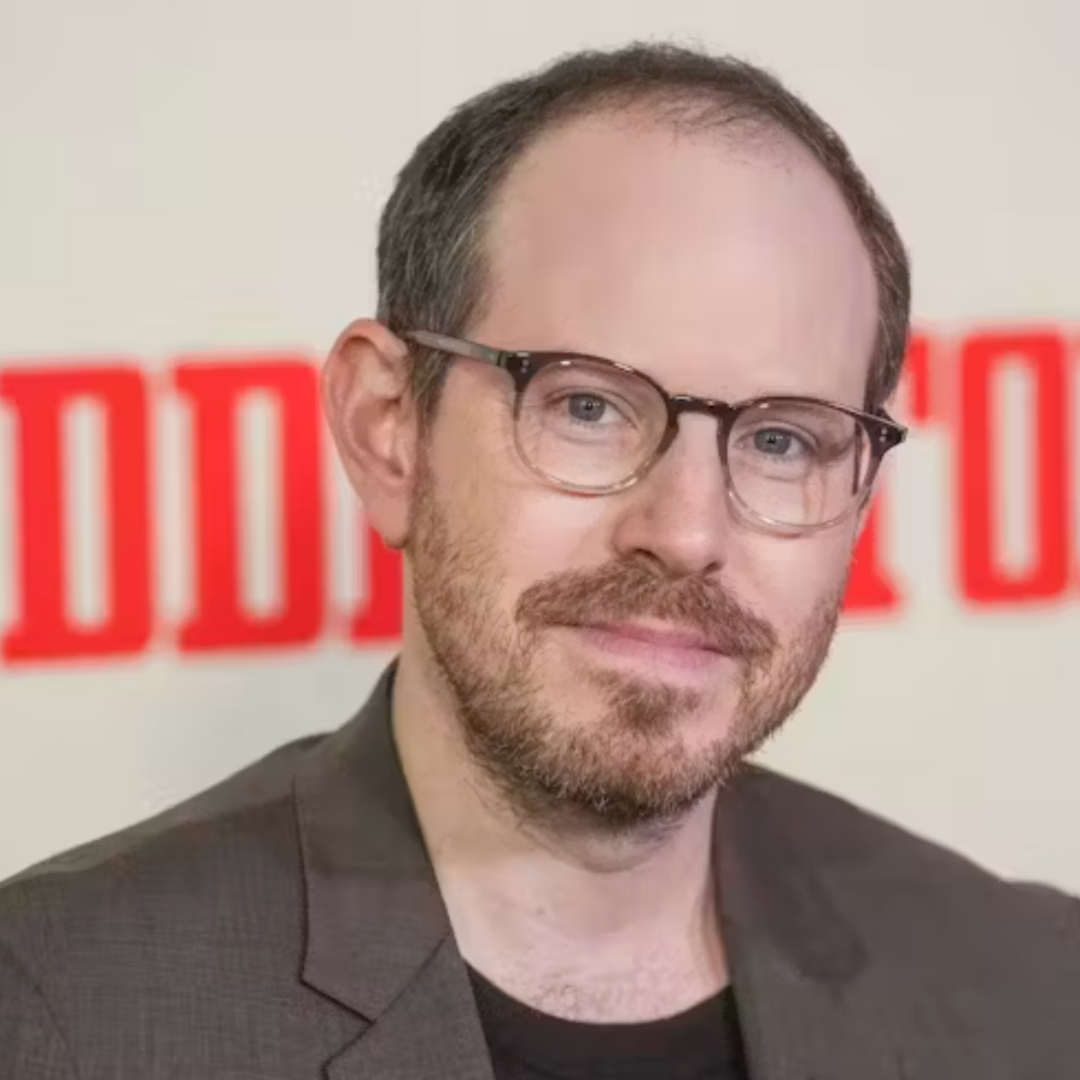 Ari Aster wearing glasses and a brown suit jacket