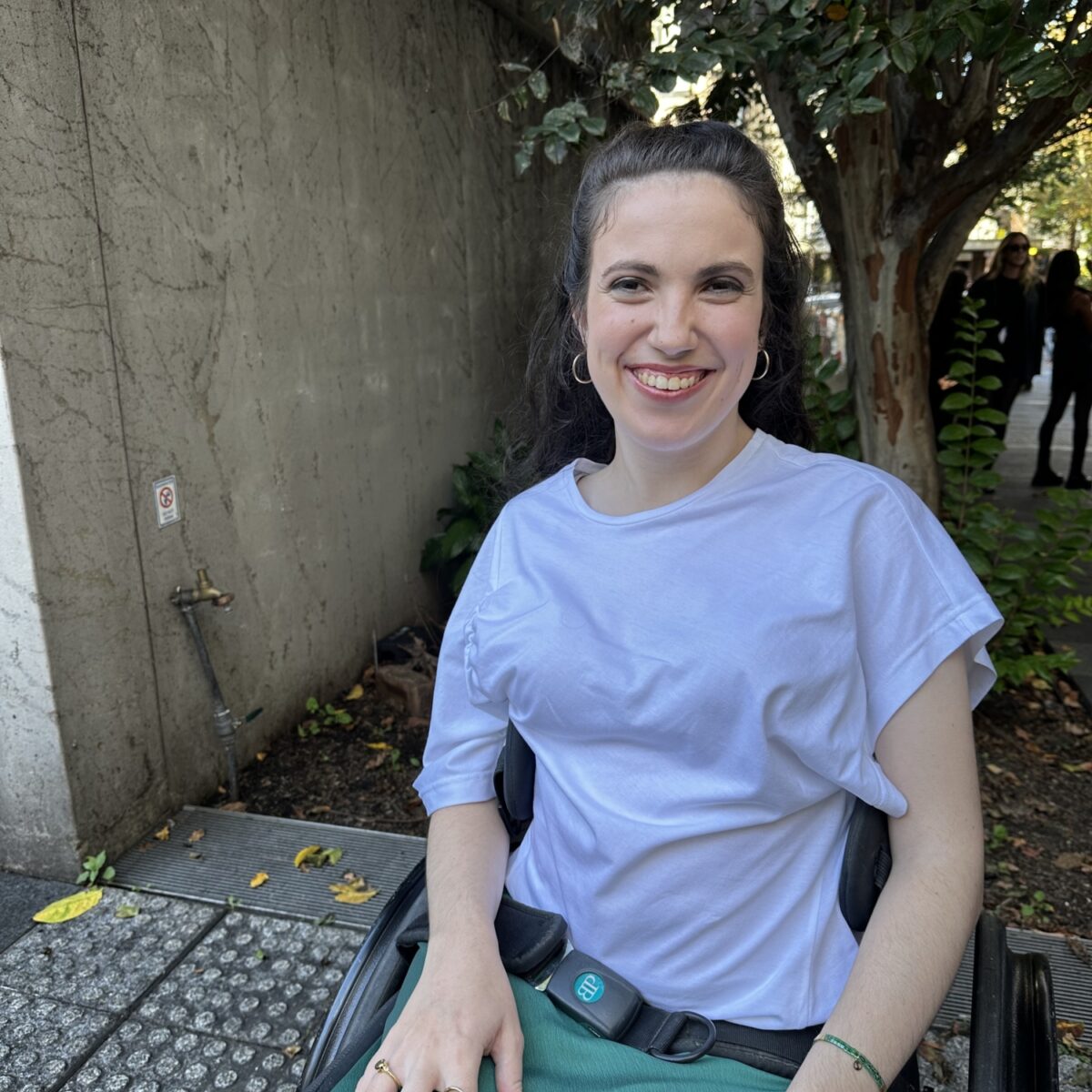 Hannah Diviney smiles at the camera. She has long dark hair, is wearing a light blue shirt, green skirt, and sitting in a wheelchair.