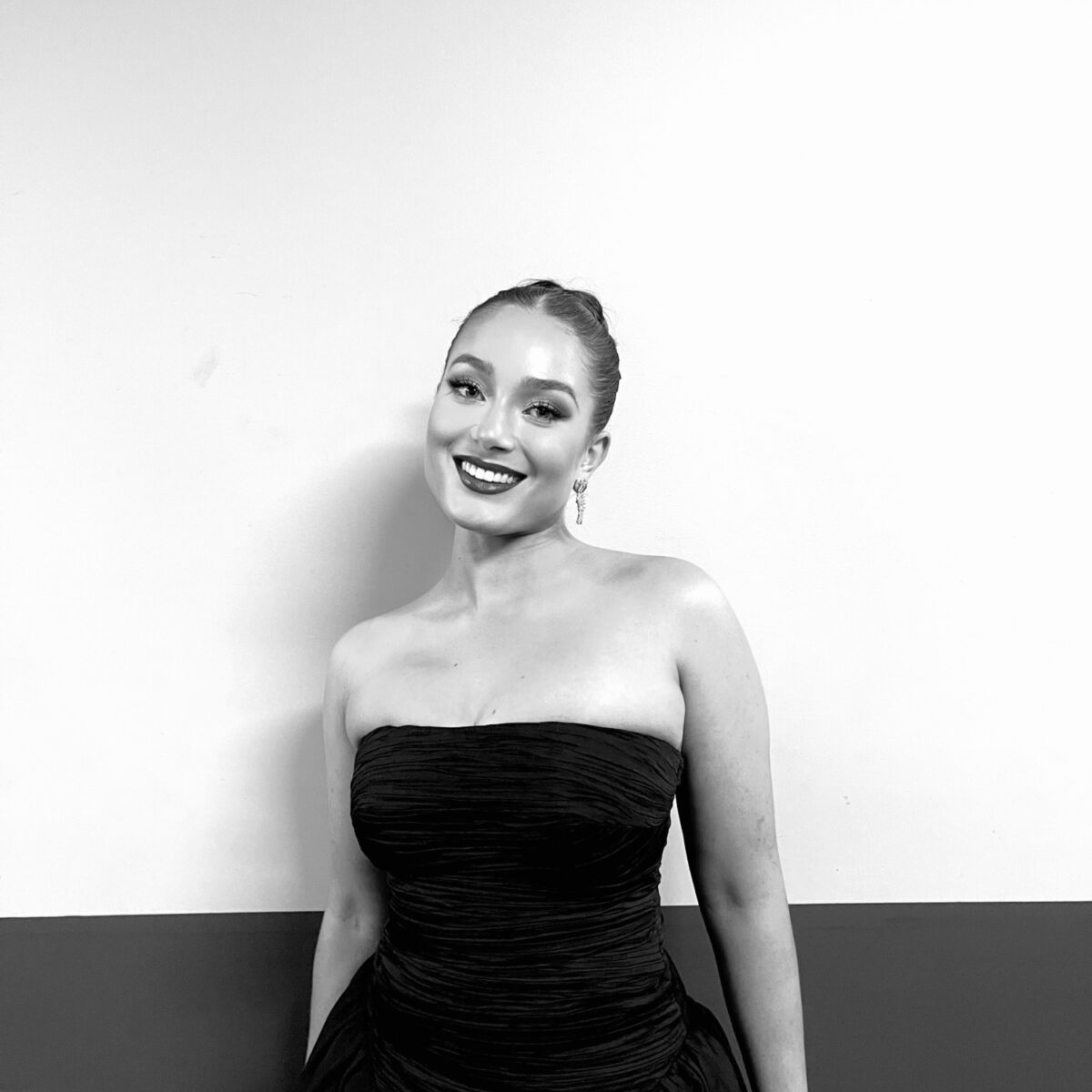 Black and white image of Emma Tomlinson she is standing smiling at the camera. She is wearing a black dress with her arms beside her body.