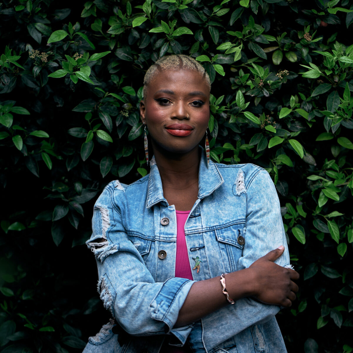 Khadija is smiling and standing in front of a leafy bush, with short blonde hair, wearing a denim jacket and a pink top.