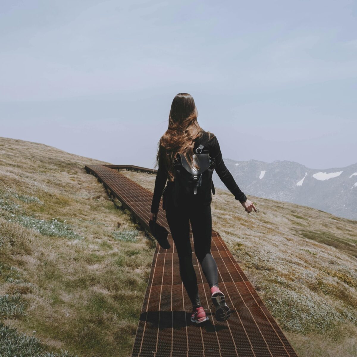 Mt Kosciuszko scene, looking from behind a woman with backpack walking along a boardwalk across sloping heathland, a snow-flecked mountain range dimly visible under grey sky in the background.