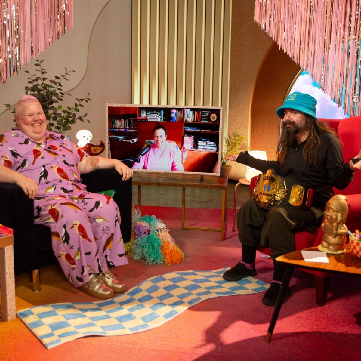 Jasper Peach is sitting in a red armchair on left side of the set of Unmasked. They are wearing a pink and bird print jumpsuit. The set is pink and sparkly and lots going on, many textures and comfy chairs. Kaya Wilson is on a tv screen in the middle of the set. On the right side of the set sits Beau Windon.