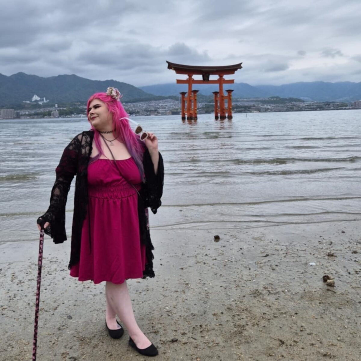 Zoe, who has pale skin, and long pink hair, wearing a long black dress with a corset belt, and lacy sleeves, while holding a black and pink floral walking stick on a beach in Japan